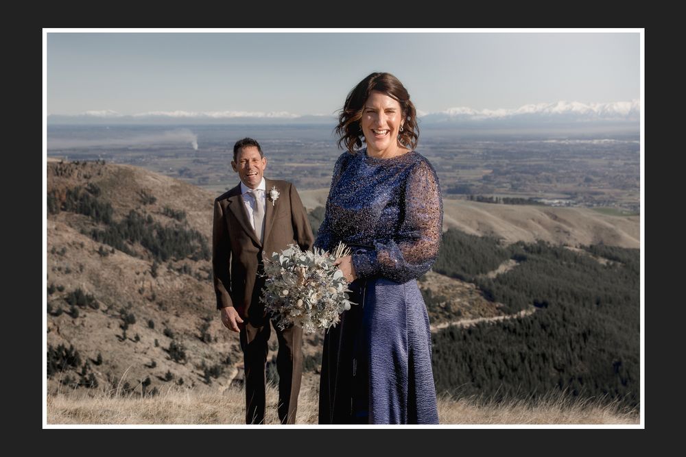 Couple poses on mountain top, bride in navy blue lace gown holding bouquet, groom in brown suit, scenic valley backdrop.