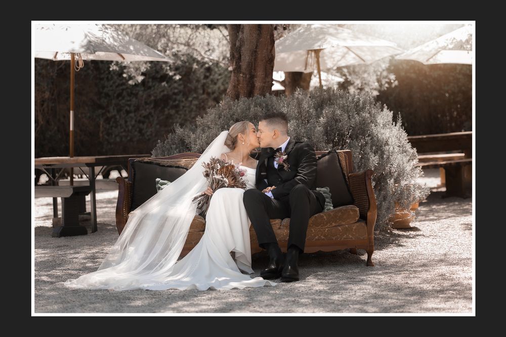 A romantic couple sits together on a vintage brown leather couch in an outdoor setting during a wedding photoshoot.