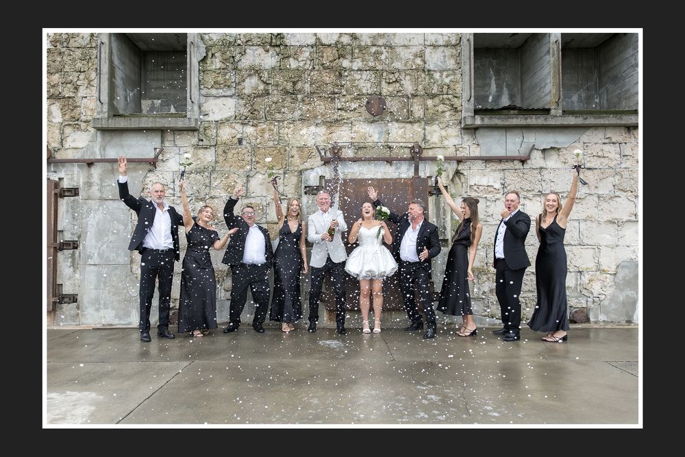 Wedding party celebrating outdoors, tossing confetti in front of a stone building.