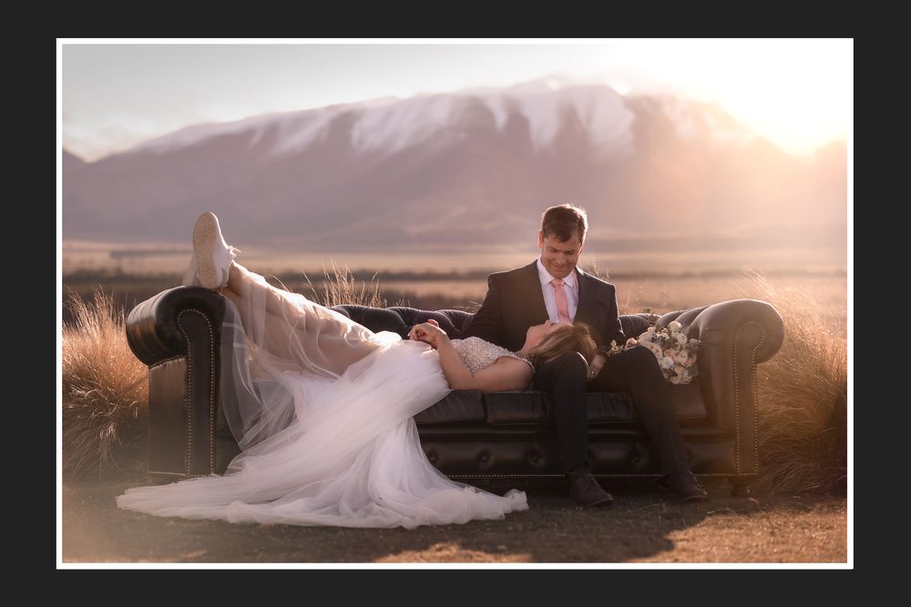 Bride and groom sit on vintage sofa at golden hour with snow-capped mountains backdrop in scenic outdoor wedding portrait.