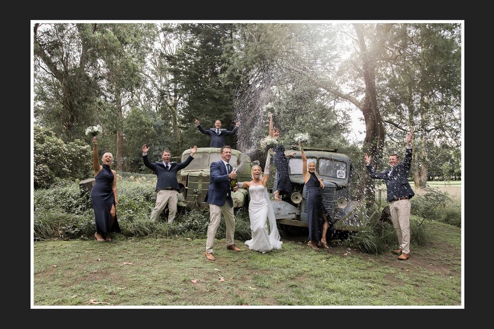 A wedding party poses outdoors near a vintage car surrounded by trees and natural greenery in a rustic setting.