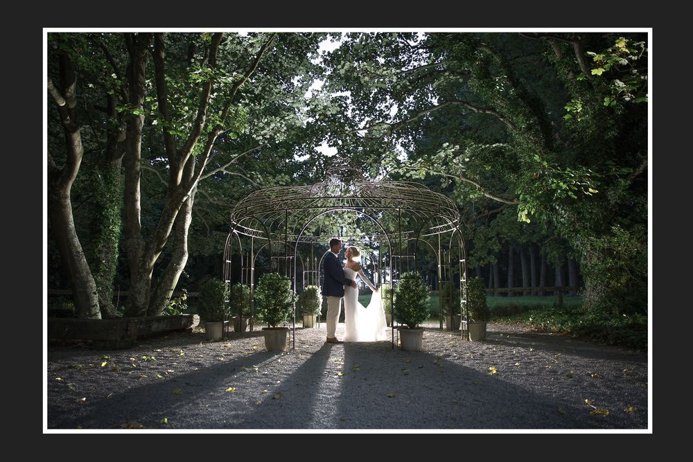 Silhouetted couple stands under an ornate garden archway surrounded by lush trees on a sunlit path.