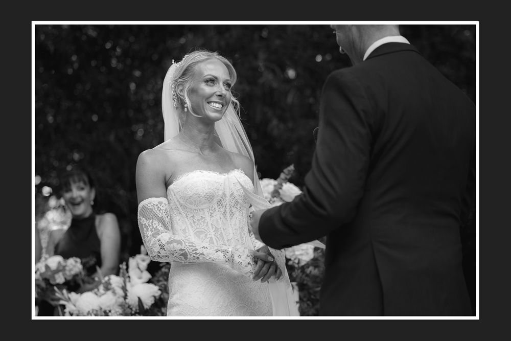 Black and white photo of a smiling bride in a strapless lace gown and veil during an outdoor wedding ceremony.