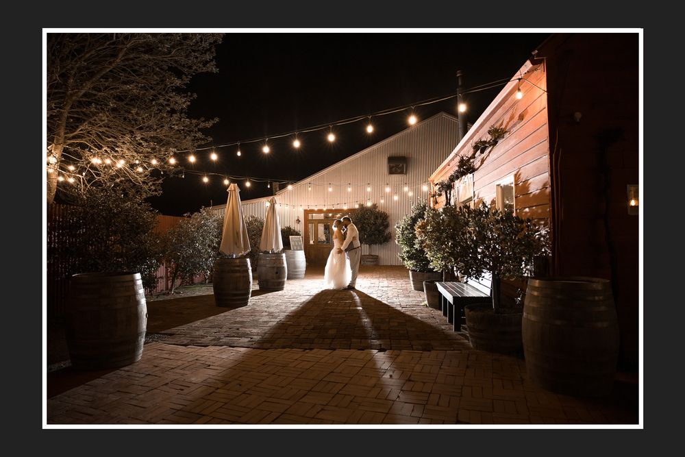 Couple in wedding attire stands under string lights at a romantic outdoor barn venue at night.