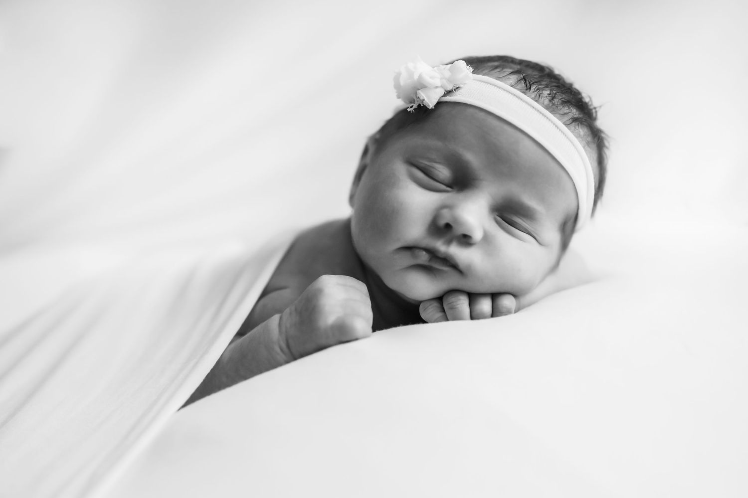 Classic black and white image of a newborn baby girl resting her head on her hands in a simple white headband.