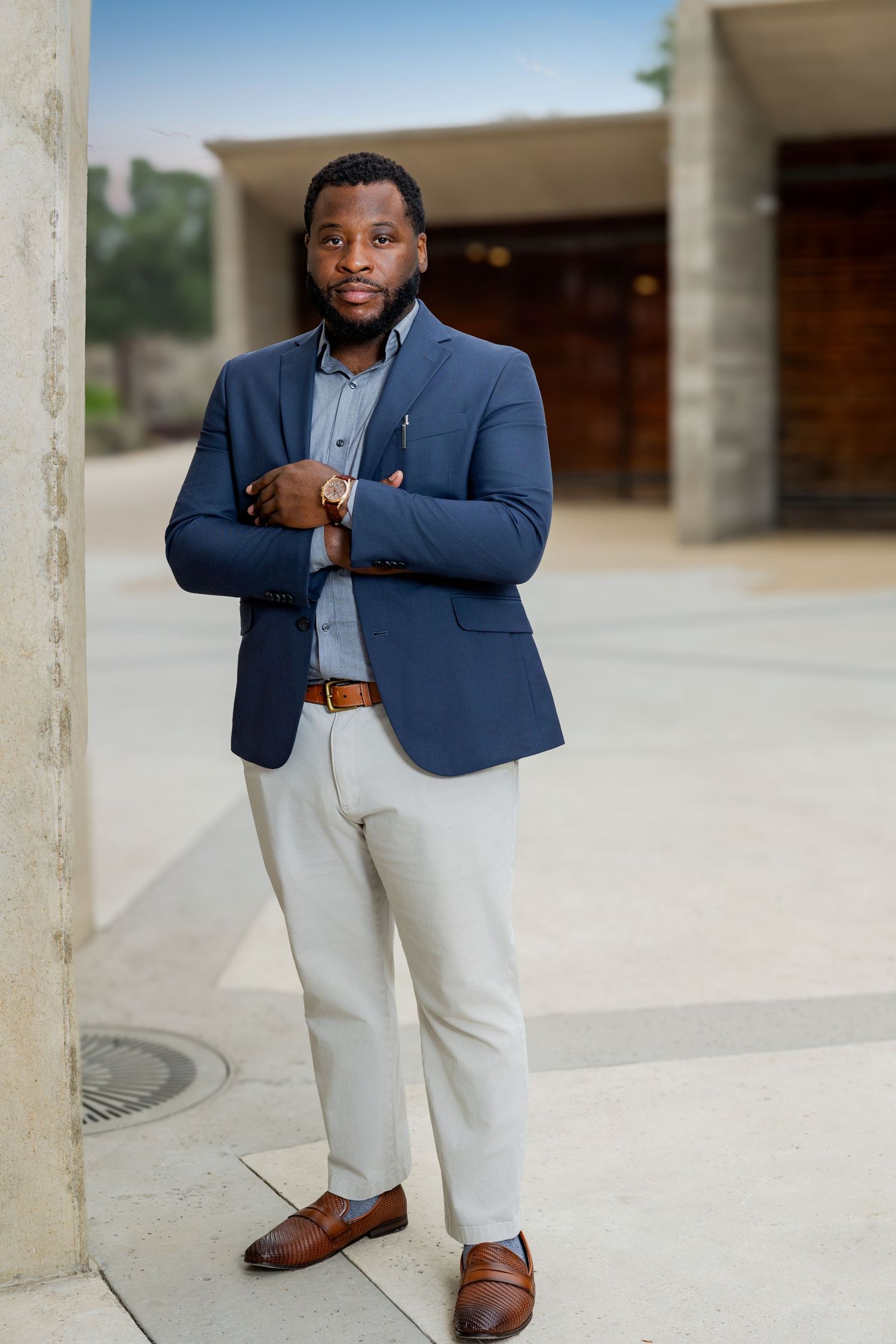 A well-dressed Black man in a navy blazer and white trousers leans against a column outdoors.