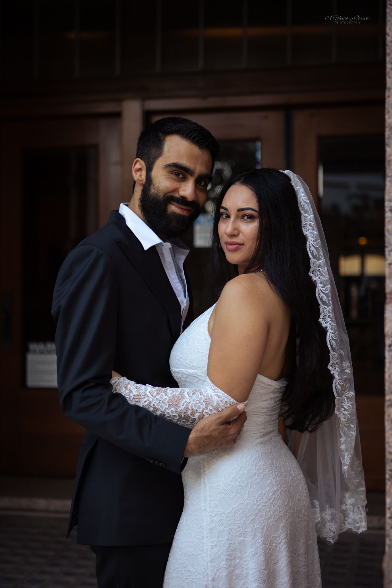 A bearded groom in a black suit embraces his bride in a white lace dress and veil, posing elegantly outdoors.