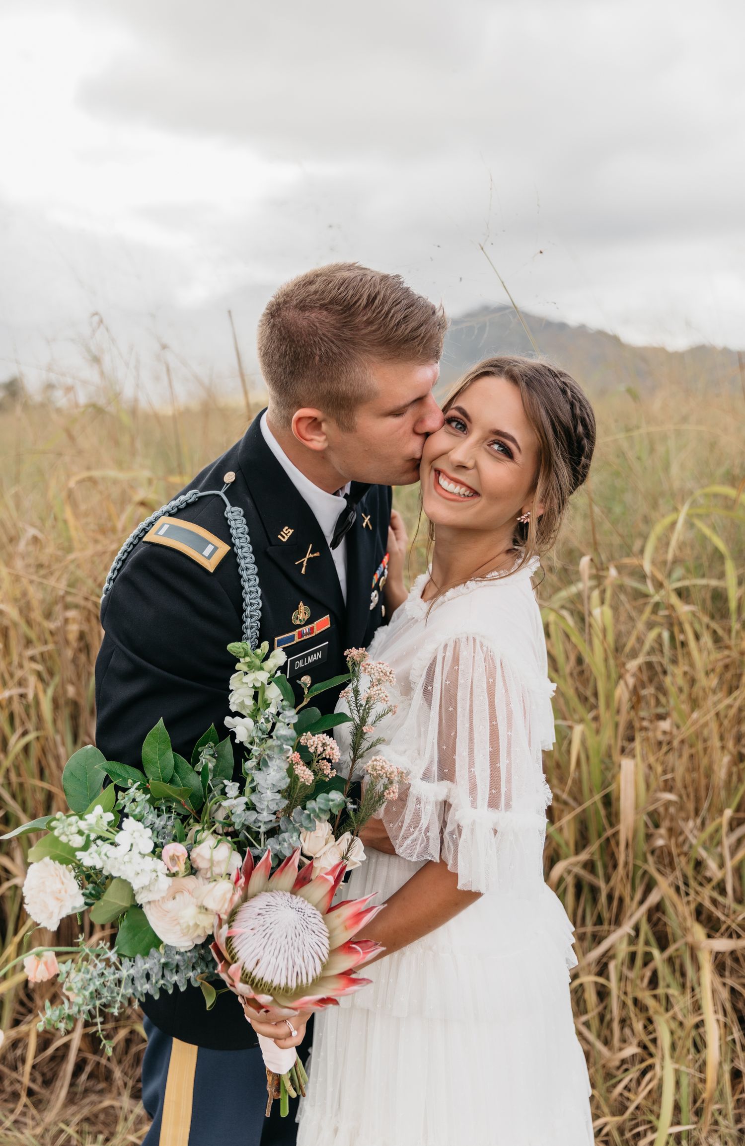 A romantic newlywed couple share intimate moments in a golden wheat field while holding a delicate floral bouquet.