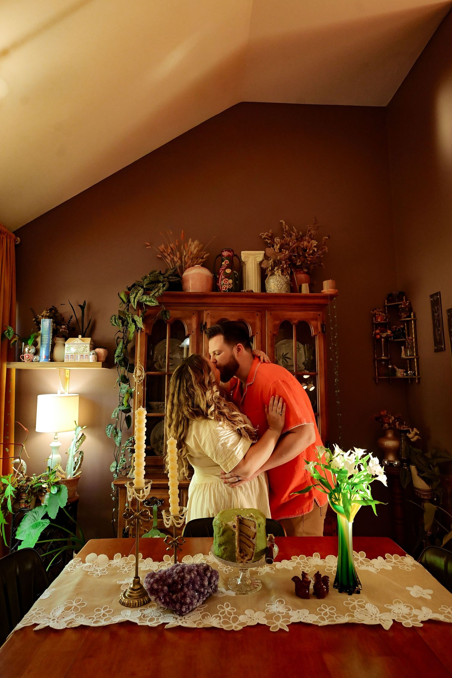 Couple sharing a romantic kiss in a warmly decorated dining room with candles, flowers, and wooden furniture.