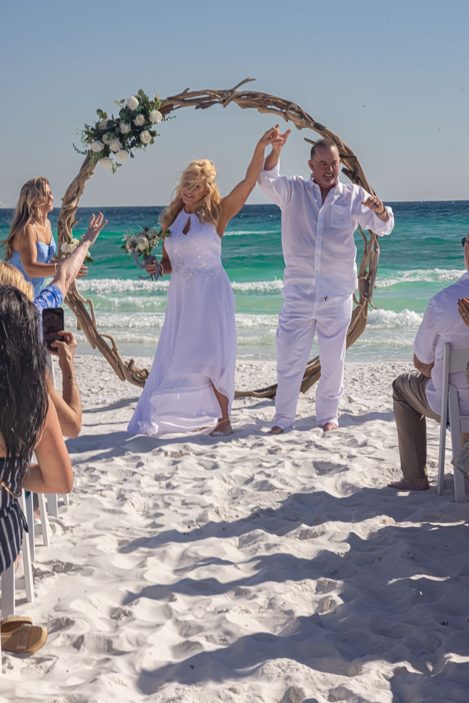 A circular wedding arch adorned with flowers frames a beach ceremony at golden hour.