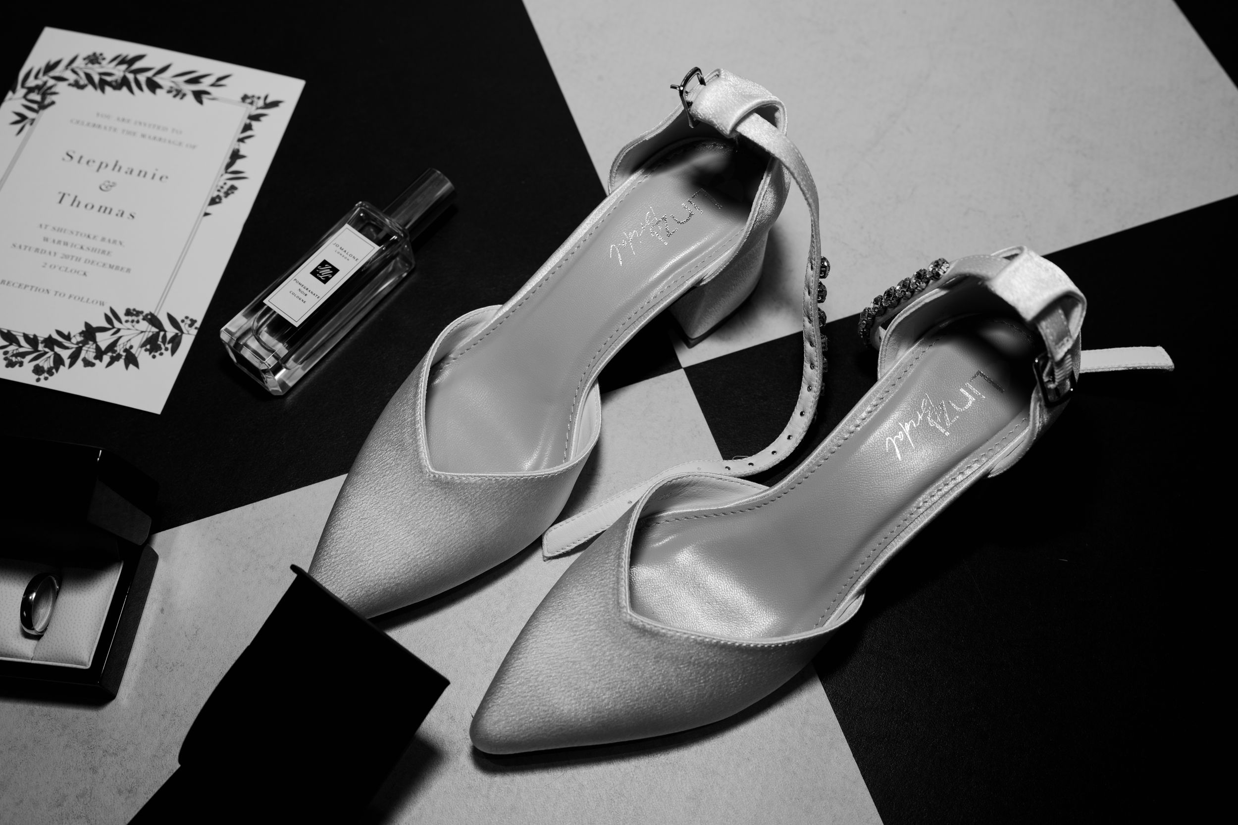 Black and white photo of elegant bridal slingback heels with bow detail, displayed with wedding invitation and Jo Malone perfume.