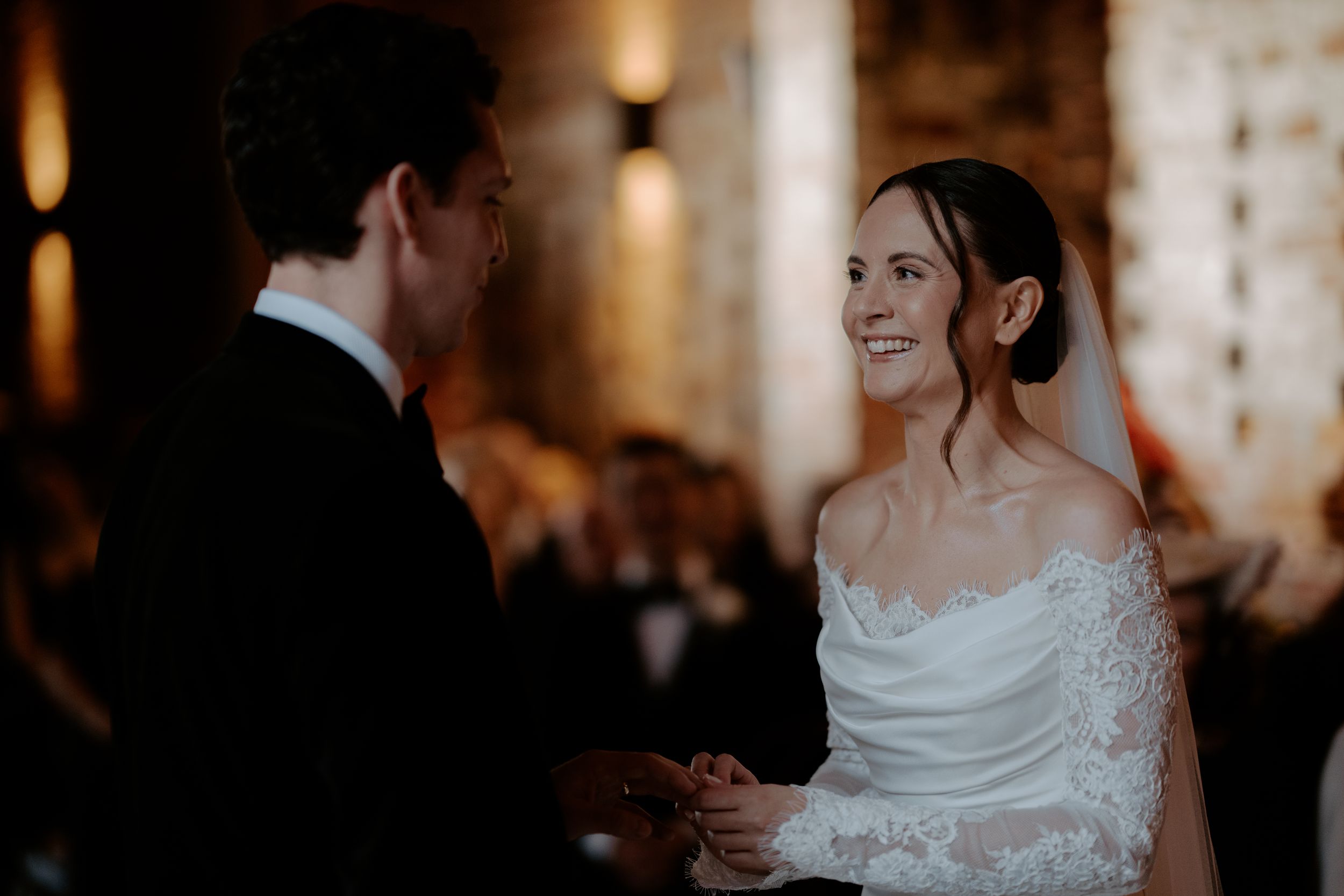 Smiling bride in lace off-shoulder gown holding hands with groom during wedding ceremony.