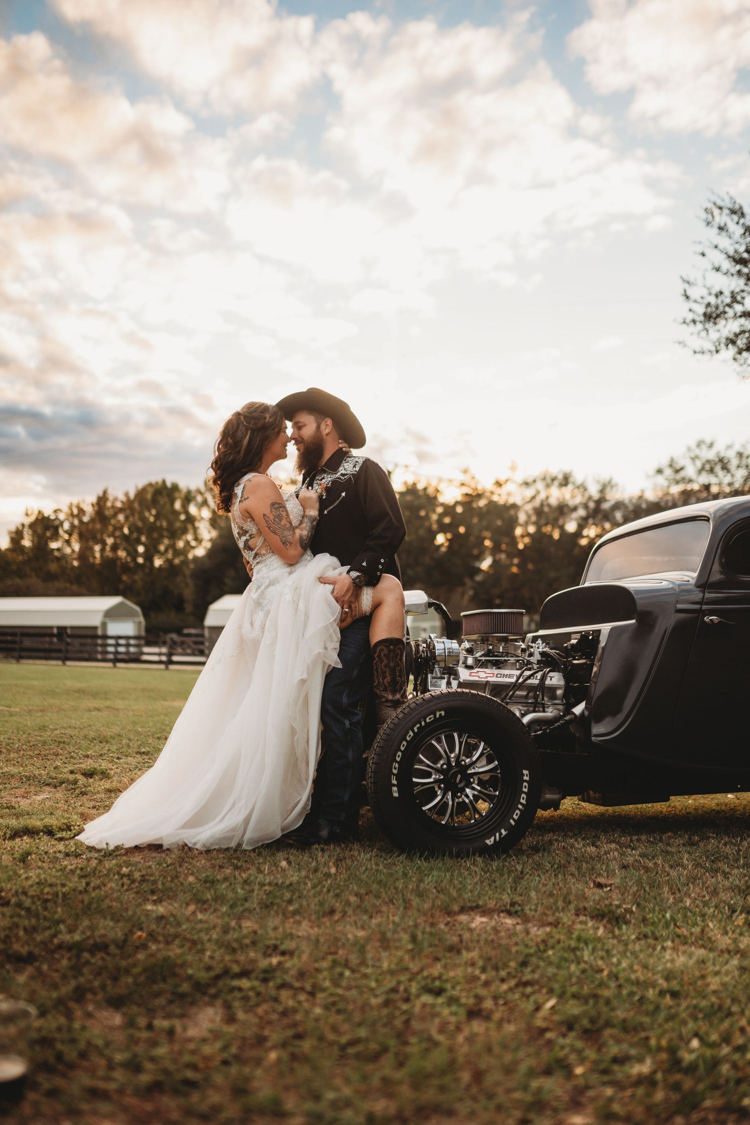 A couple in wedding attire poses by a vintage car at sunset.
