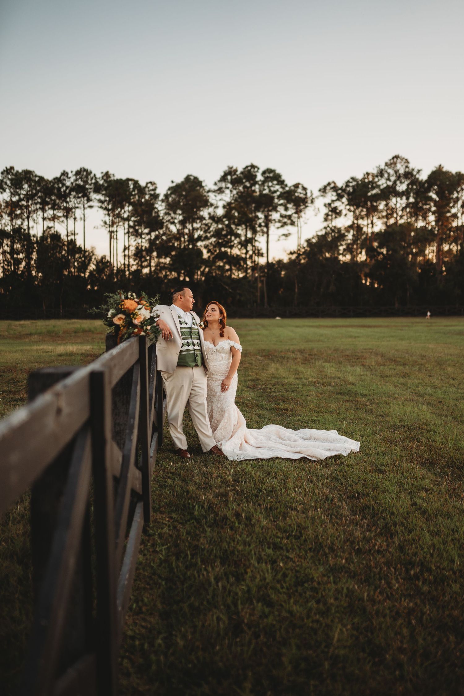 Sunset wedding portrait by wooden fence with tall pine trees in background.