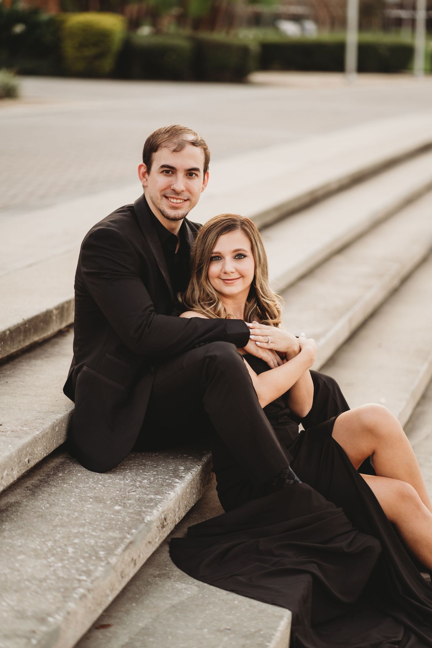 Couple in black attire sitting casually on concrete steps during engagement photoshoot.