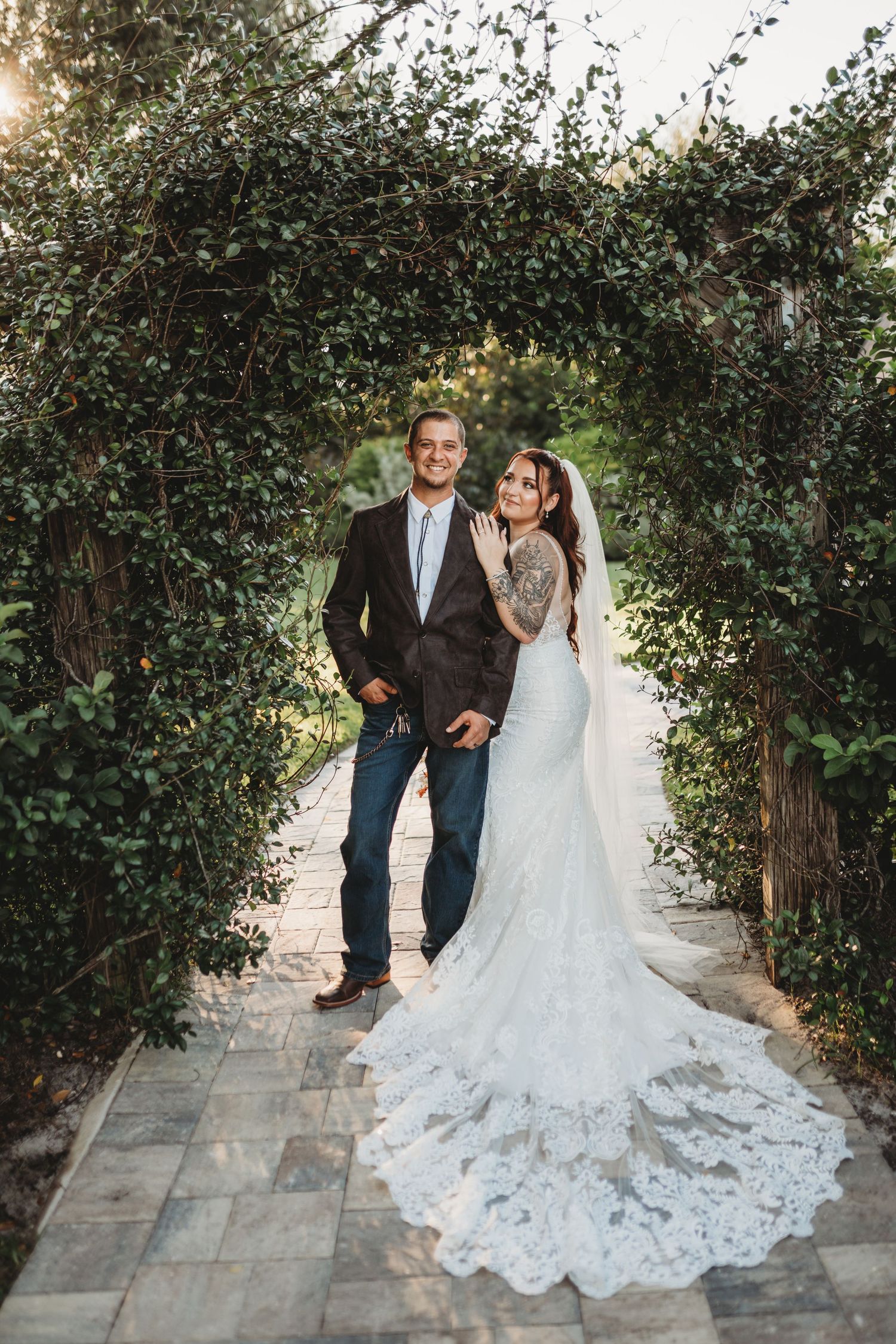 Bride in lace gown and groom pose under garden archway on stone path.