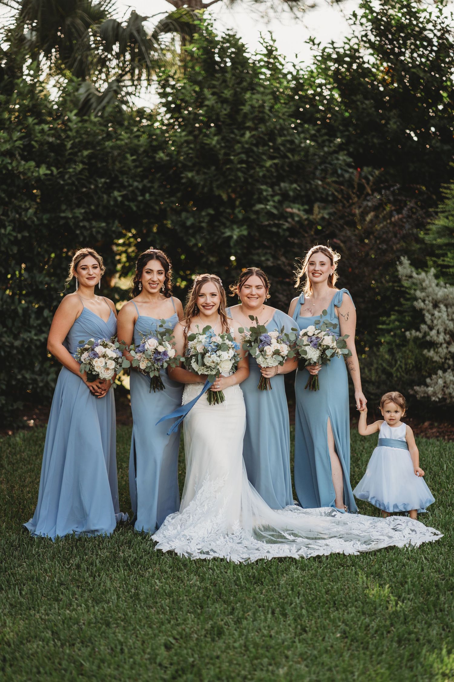 Bridal party in light blue dresses poses on lawn with trailing white wedding gown.