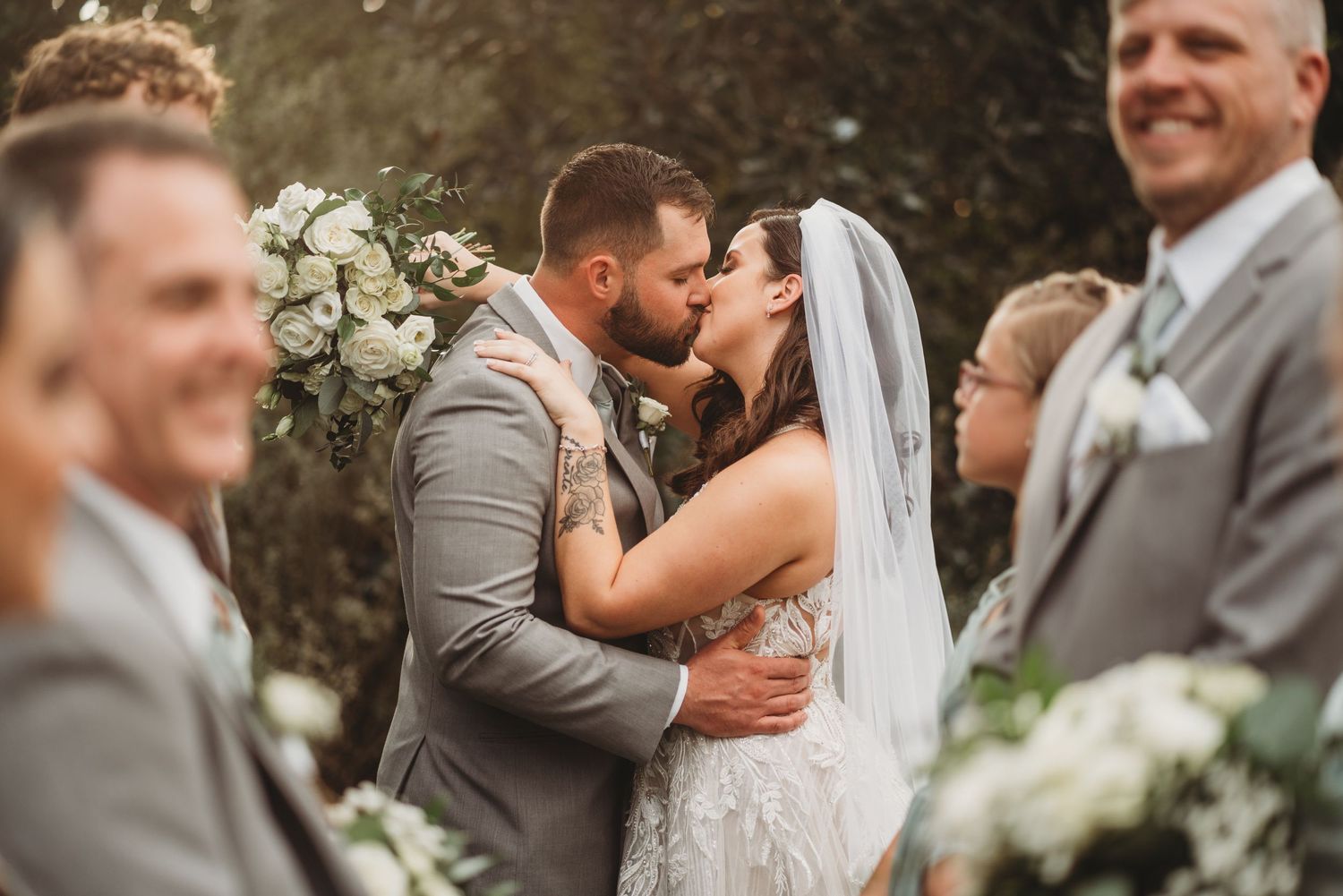 Romantic first kiss moment during outdoor wedding ceremony with guests looking on.