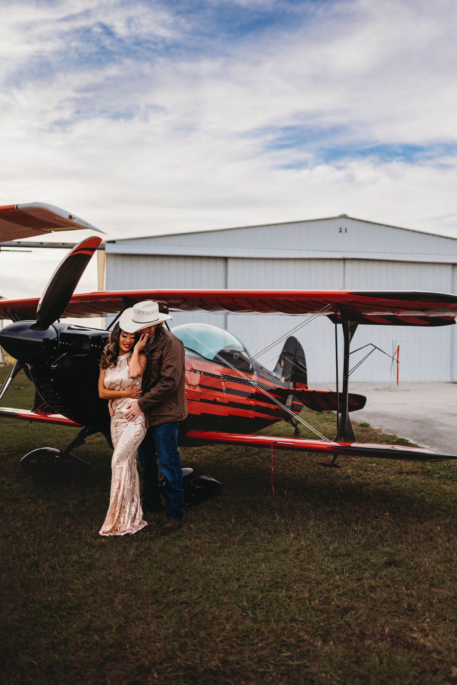 A couple embraces in front of a vintage red and white airplane with hangar in background.