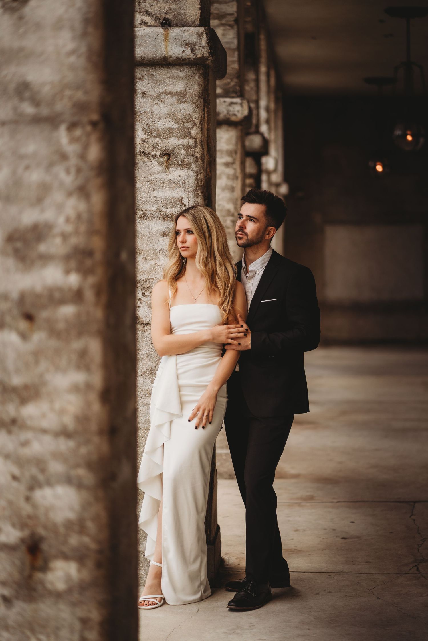 Elegant couple in formal attire pose along stone colonnade hallway.