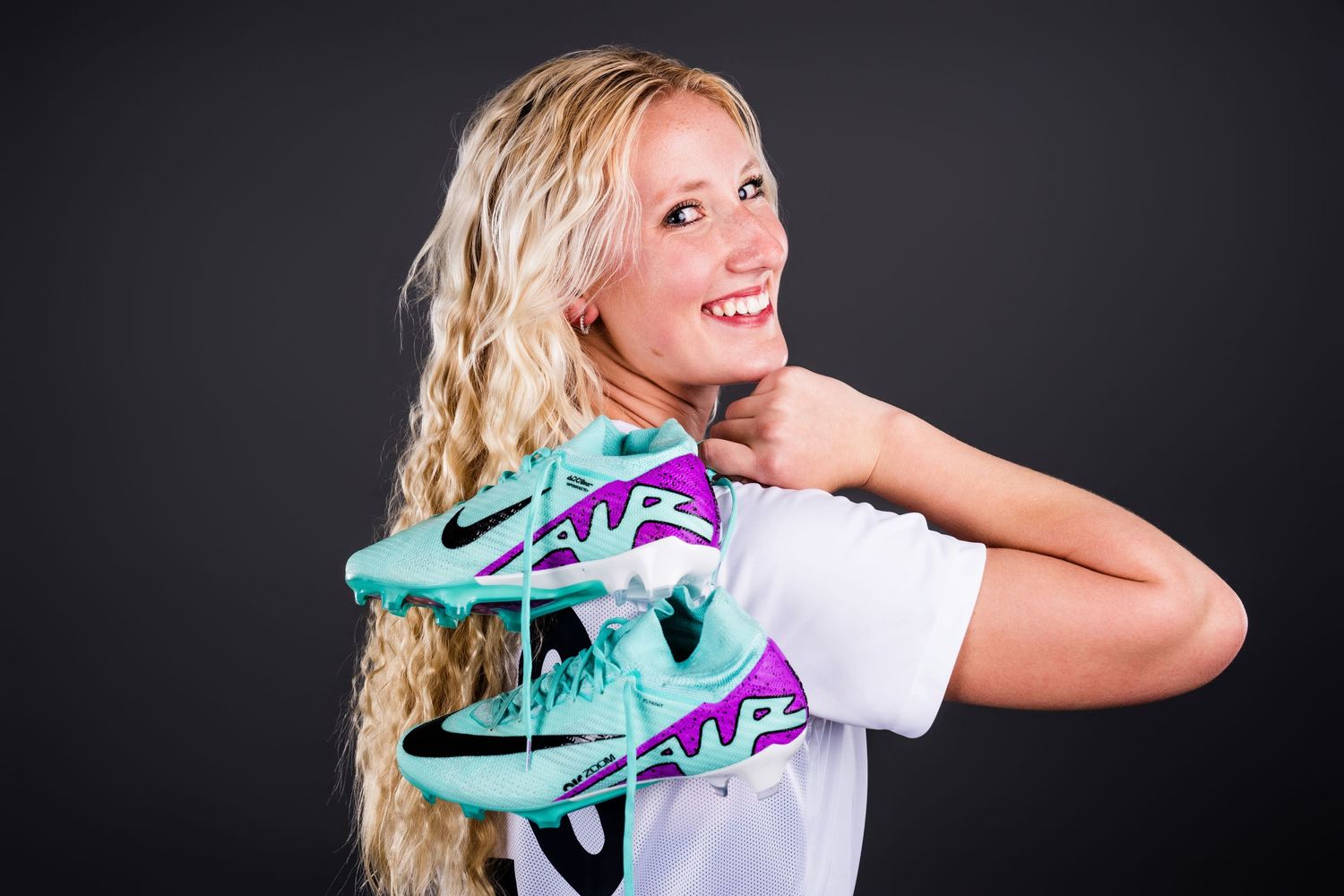 Person with long blonde curly hair holds teal and purple roller skates against dark background.