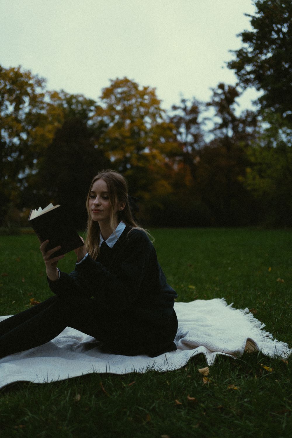 Junge Frau im Vintage-Stil mit schwarzen Rock, schwarzen Pullover und weißer Bluse liest ein Buch auf Decke im Großen Garten in Dresden