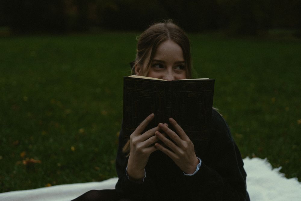 Portrait Fotoshooting einer schmunzelnden Frau, die sich mit Gesicht hinter Buch halb versteckt im Großen Garten in Dresden