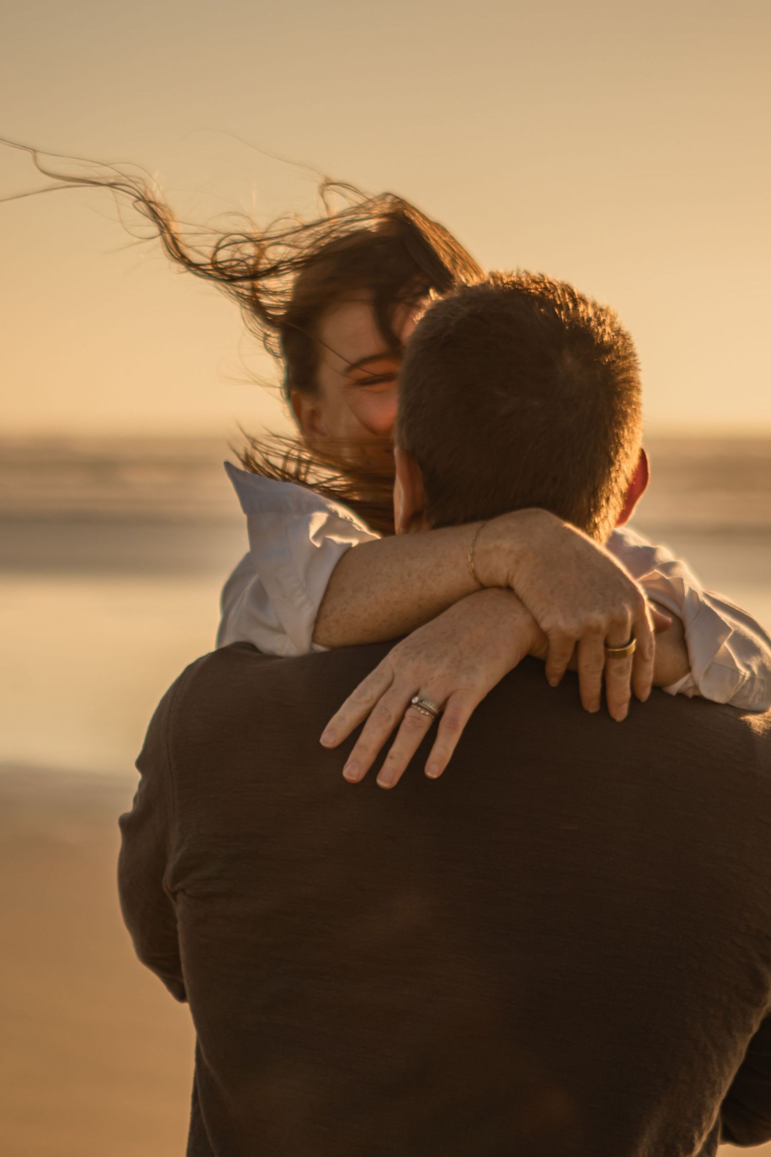 A couple hold each other playfully on the beach. He is swinging her in the wind.