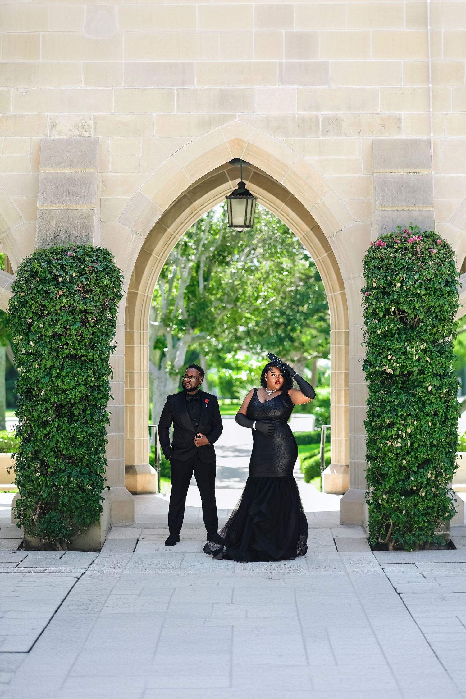 Couple in matching black formal attire posing under stone Gothic arch surrounded by lush greenery.