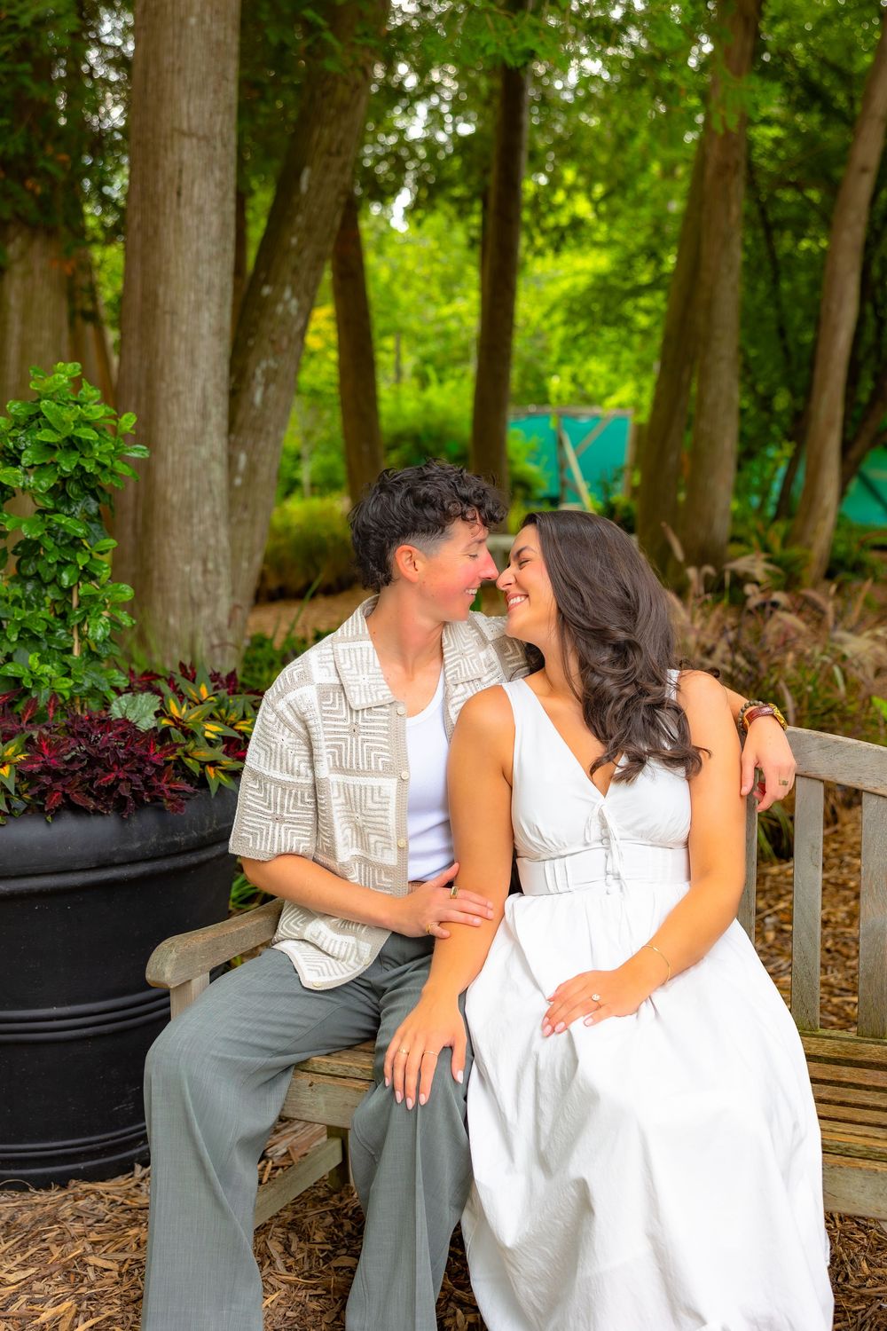 A couple shares an intimate moment while sitting on a wooden bench in a garden setting with tall trees.