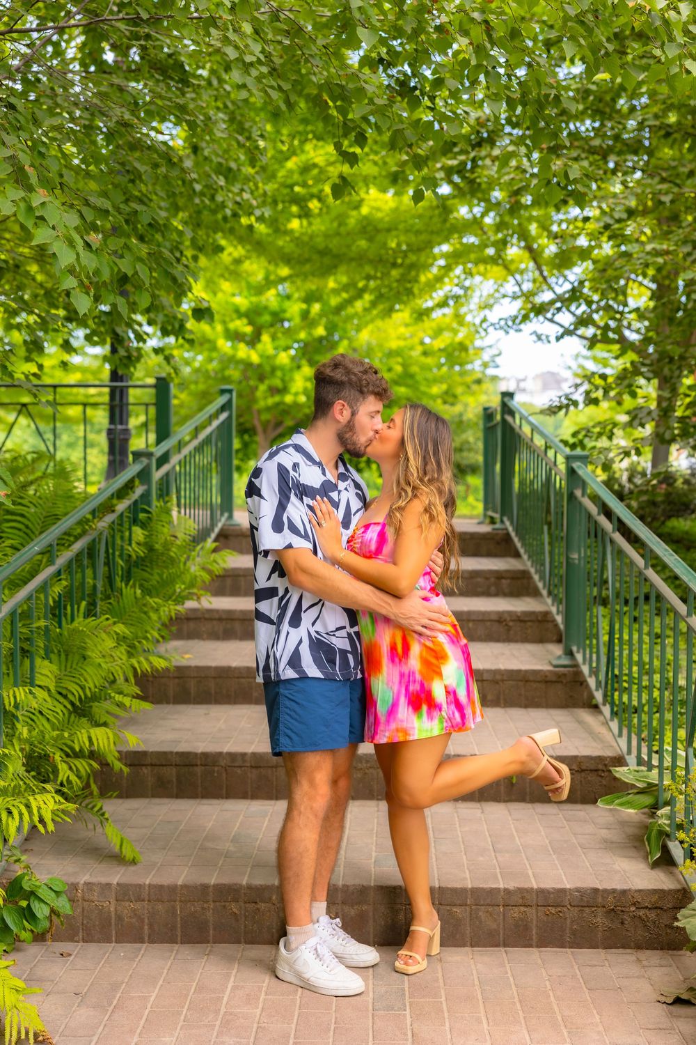 A couple in colorful summer outfits share a romantic moment on outdoor stairs surrounded by lush green foliage.
