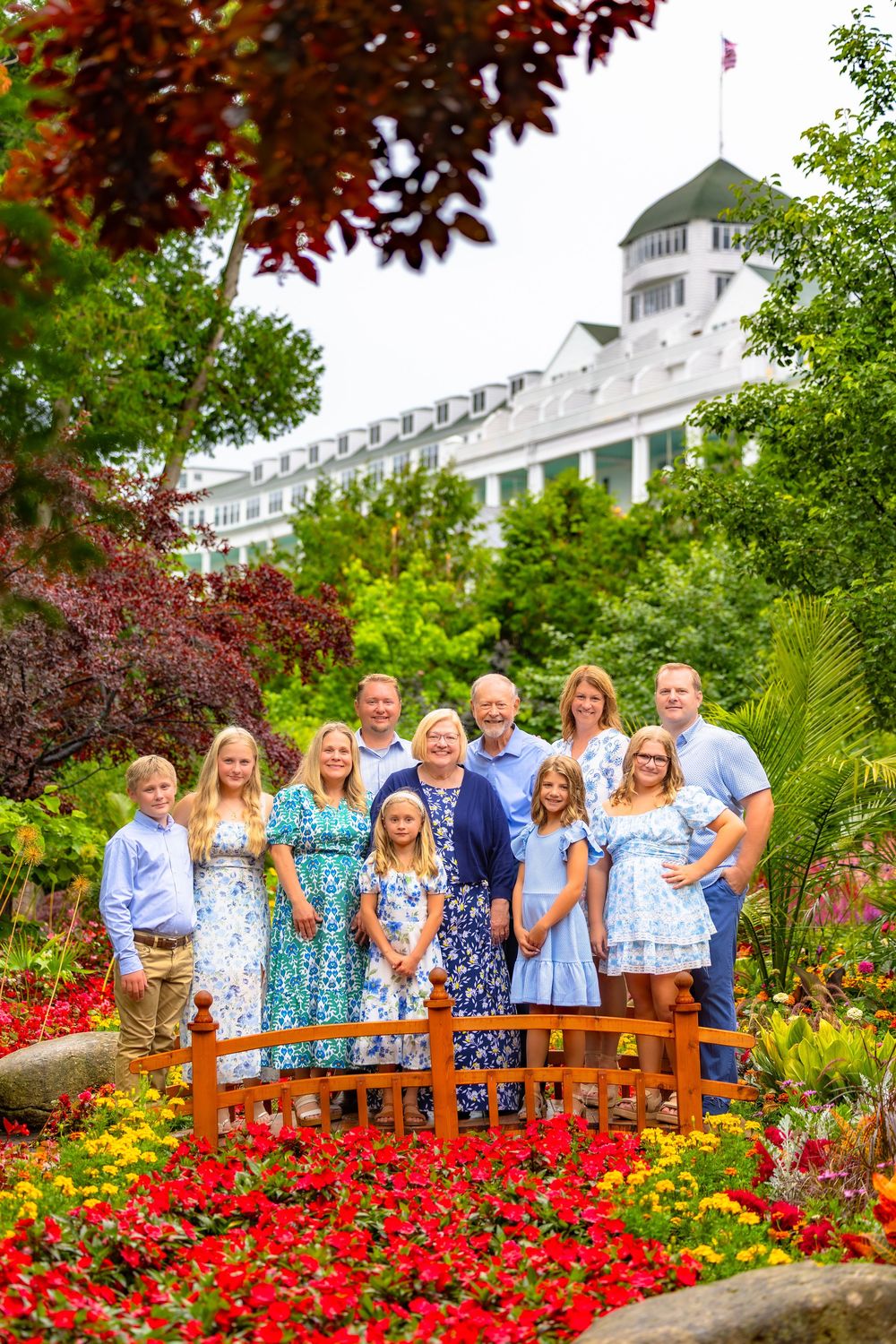 A group stands on a wooden bridge in a vibrant garden with red flowers and a white building in the background.