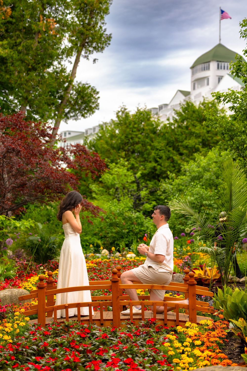 A romantic proposal moment unfolds on a wooden bridge surrounded by vibrant garden flowers and greenery.