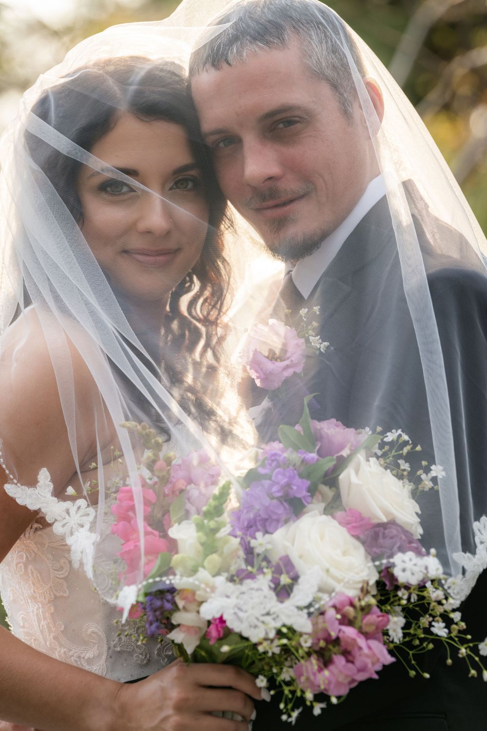 A bride and groom pose together under a veil with pink and purple floral bouquet on their wedding day.