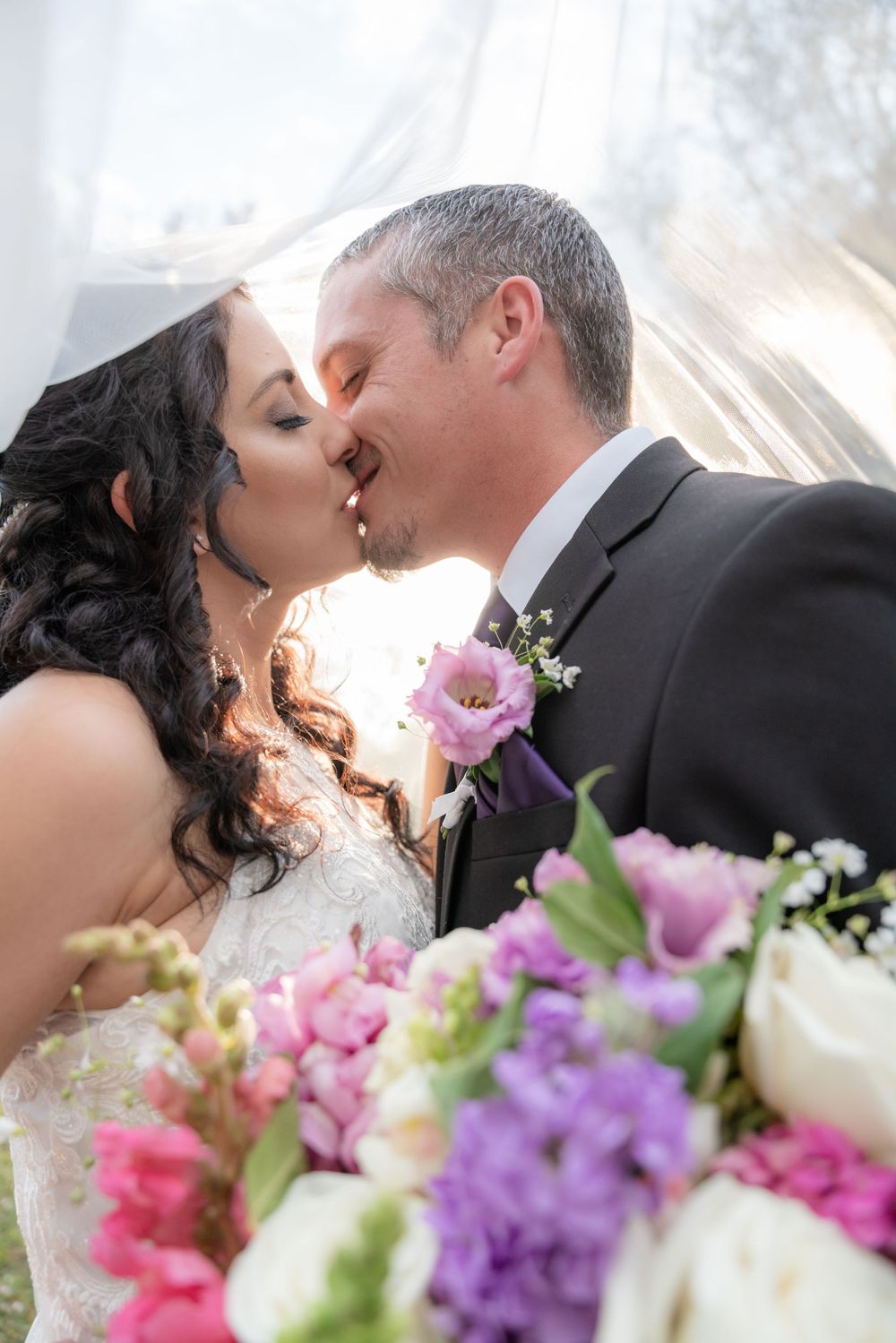 Romantic wedding kiss captured with beautiful bouquet of pink and purple flowers in the foreground.