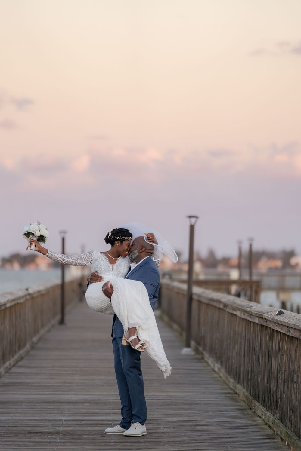 A romantic moment captures two people embracing on a wooden pier at sunset with soft pink skies in the background.