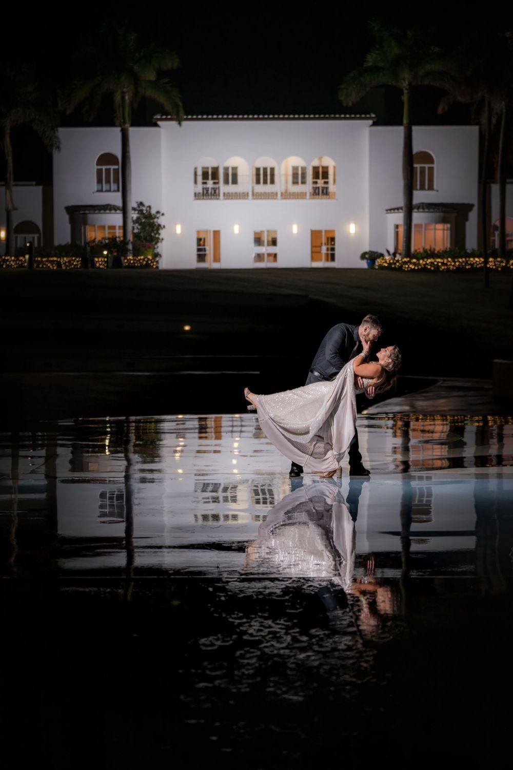 Romantic wedding couple poses at night in front of The Mansion at Tuckahoe with palm trees and reflection in water in Stuart, Florida.