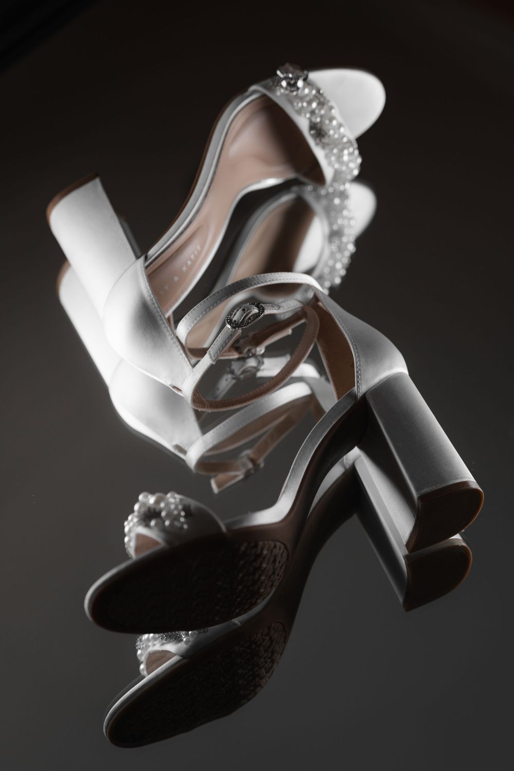 Elegant white wedding shoes with crystal embellishments and block heels arranged artistically against dark background.