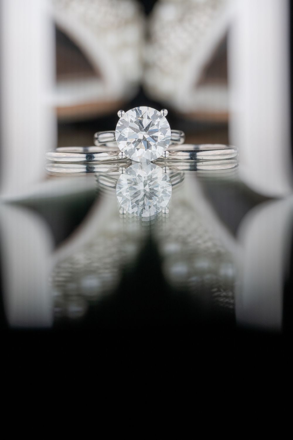 Elegant solitaire diamond engagement ring with split shank design shown with reflection on dark mirrored surface.