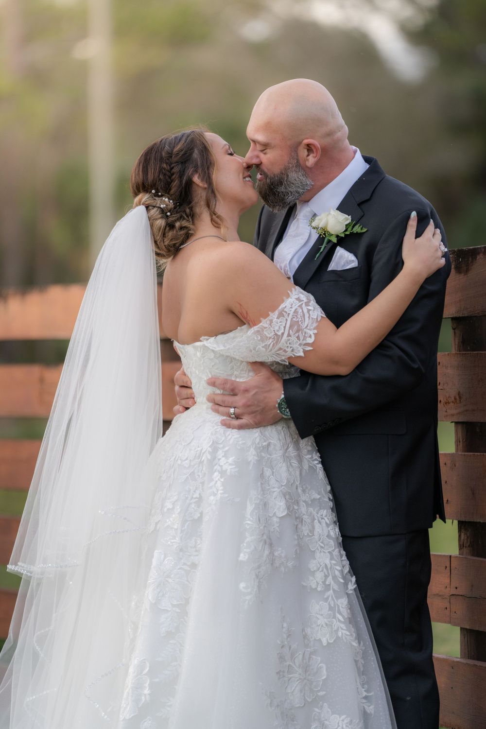 A bride in a lace wedding gown and veil embraces her groom wearing a black suit during an intimate outdoor moment.