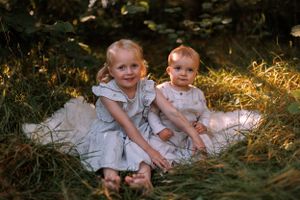 Two young children in white dresses sit together on a blanket in dappled sunlight among green foliage.