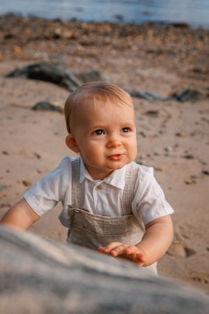 A baby in a white shirt and tan overalls sits on sandy ground during golden hour with a warm natural expression.
