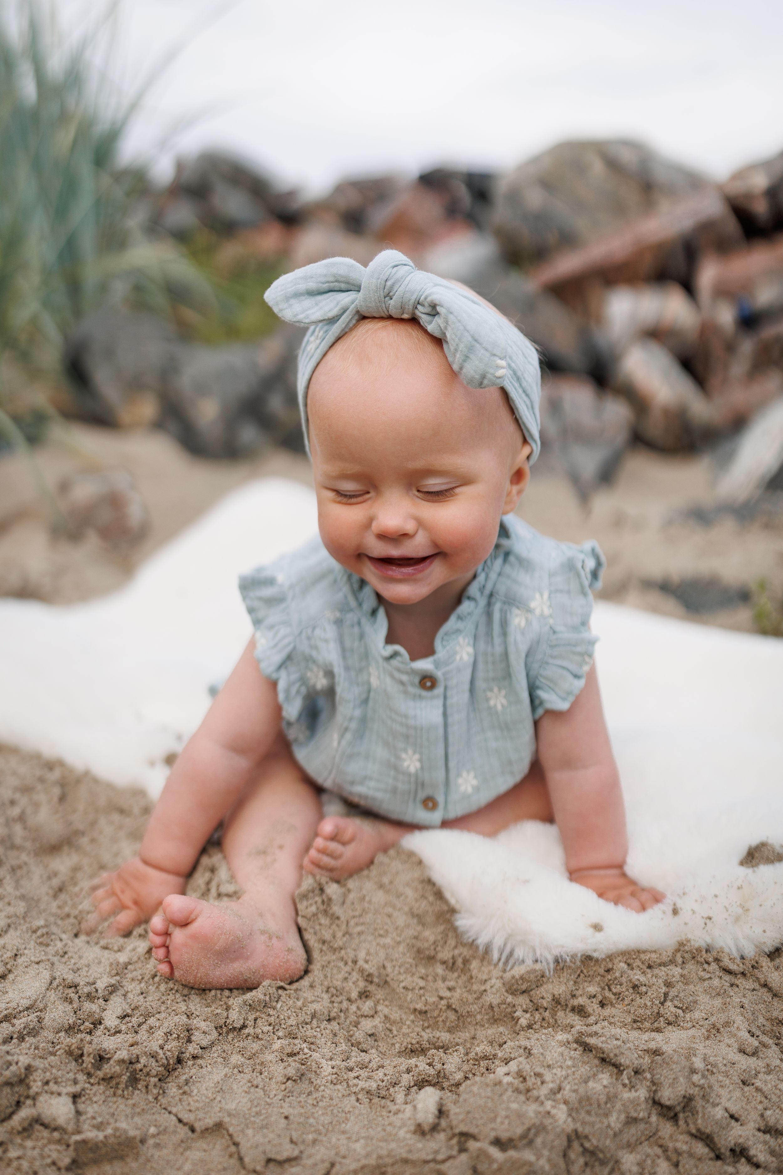Baby wearing blue ruffled top and matching headband plays in sand on sunny beach.