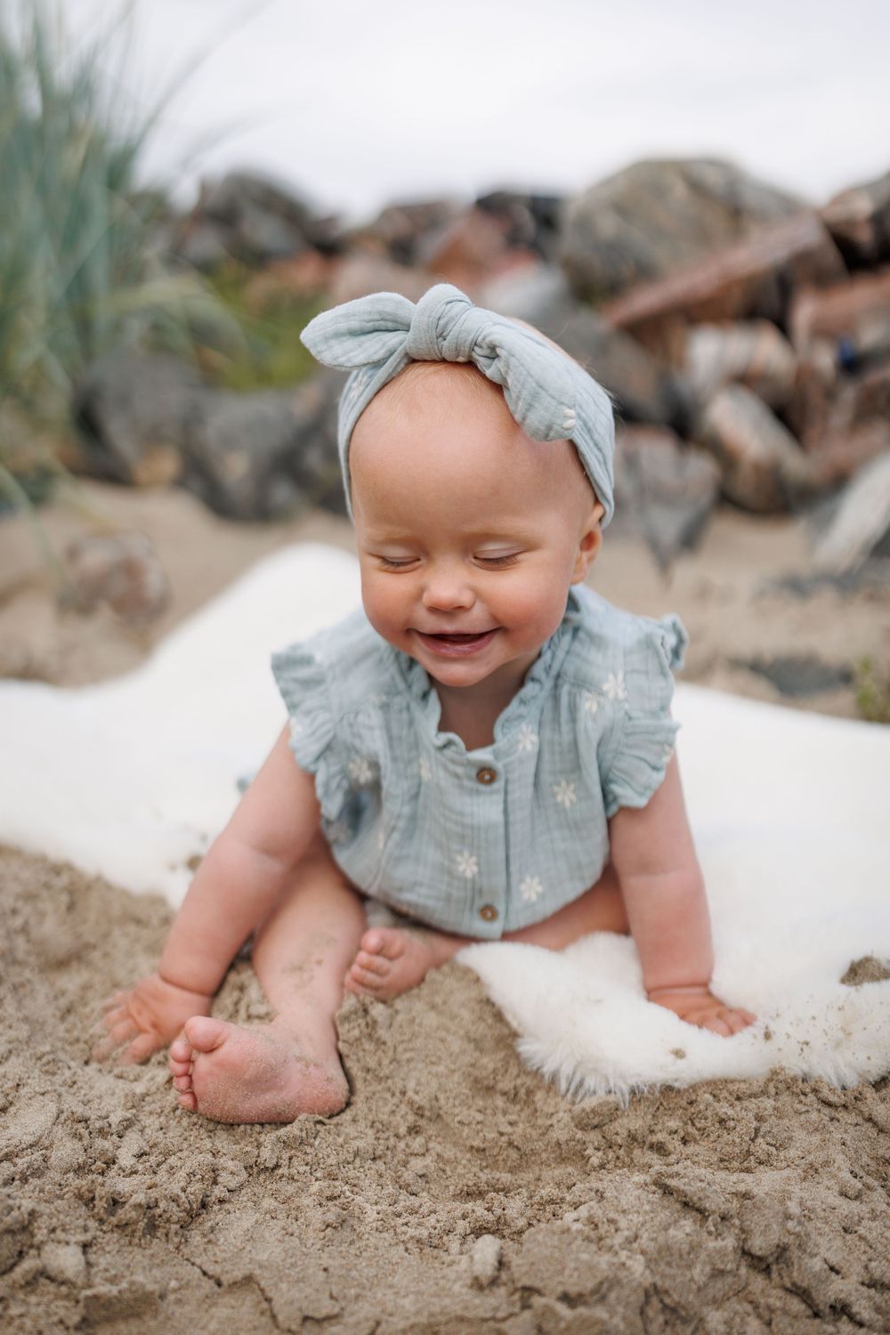 Baby wearing blue ruffled top and matching headband plays in sand on sunny beach.