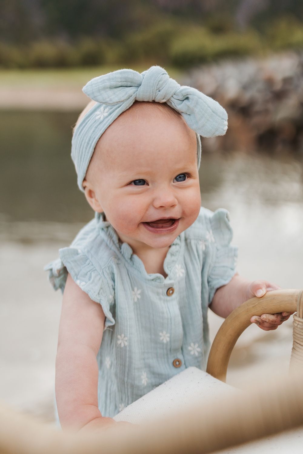 Baby in a light blue ruffled dress and matching headband smiles brightly while holding onto something outdoors.