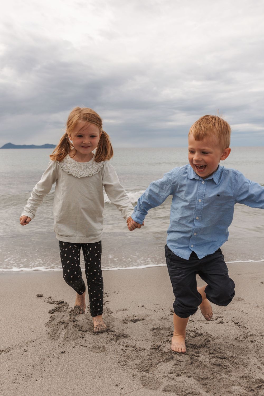 Two children holding hands and running joyfully on a beach with mountains and cloudy skies in the background.