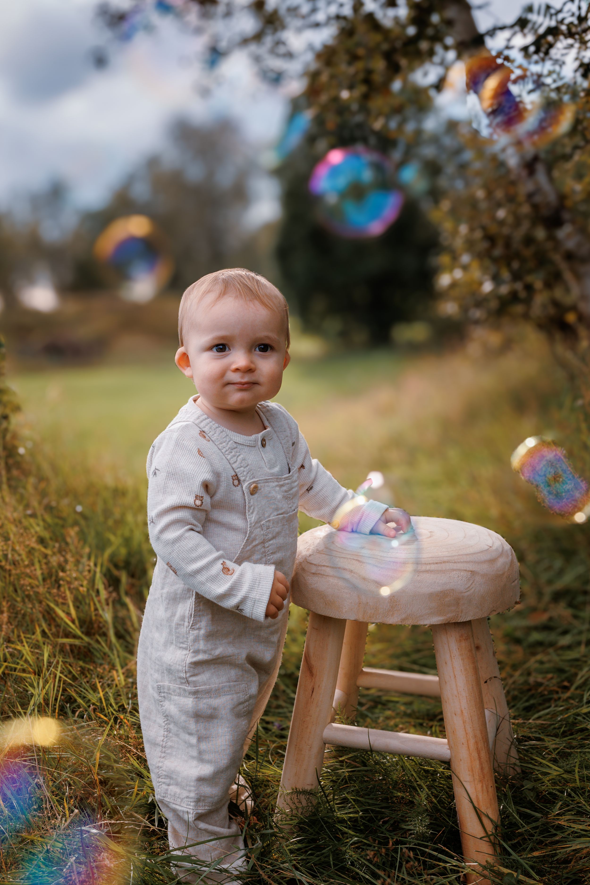 A small toddler in light colored clothing stands by a wooden stool outdoors while soap bubbles float through the air.