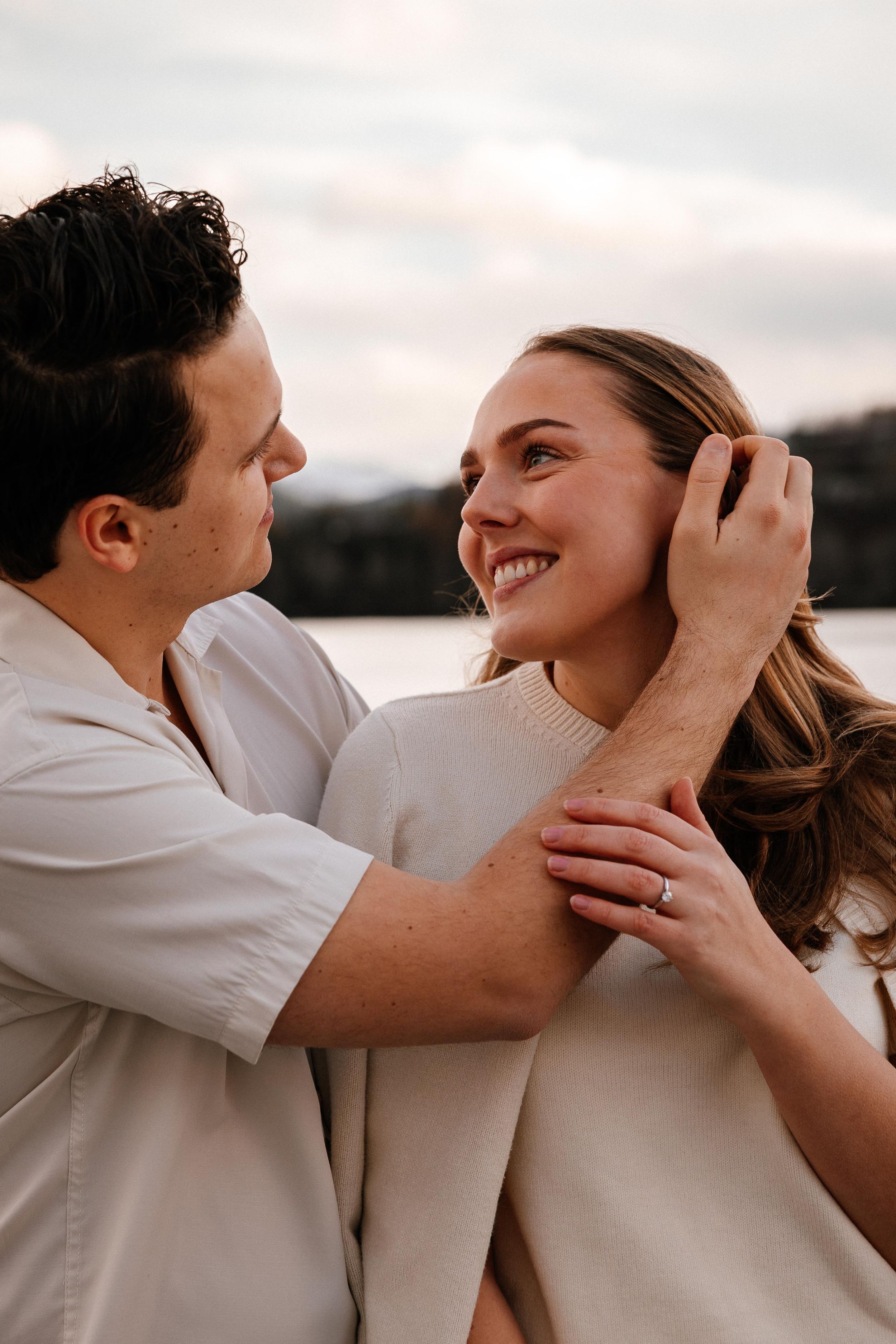 A tender romantic moment between a couple wearing neutral colors as they embrace outdoors at sunset by the water.
