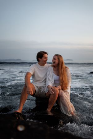 Couple sitting on rocks at beach during sunset sharing a joyful moment as waves crash around them.