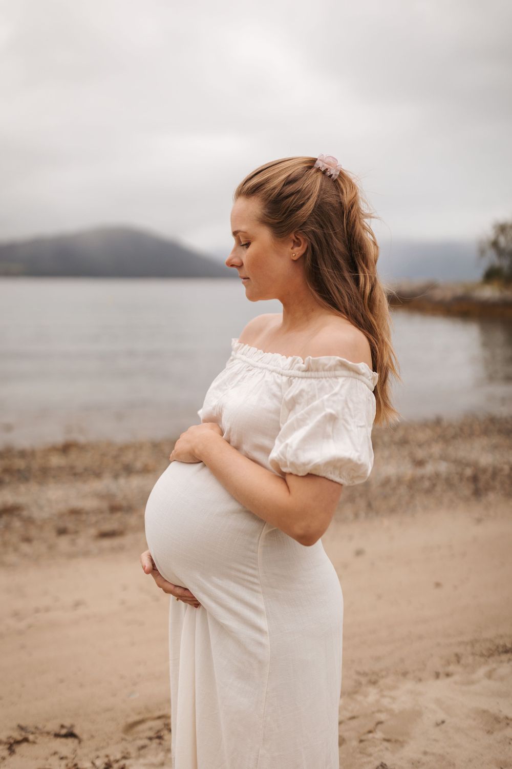 Pregnant woman in white off-shoulder dress stands thoughtfully on beach shore with mountains in misty background.
